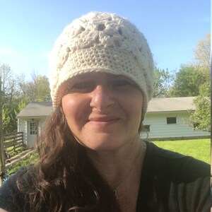 Peut inclure: Un chapeau crocheté blanc avec un motif en dentelle. Le chapeau est porté par une personne aux cheveux longs et bruns. La personne sourit et regarde la caméra.