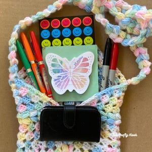 May include: A pastel-coloured crocheted bag filled with stationery items. The bag contains pens, markers, a small notebook with a butterfly design, a sheet of smiley face stickers, and a black wallet. The bag has a long strap.