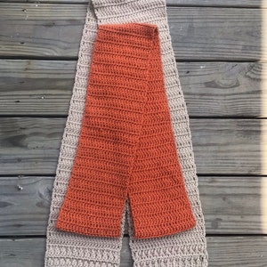 May include: A crocheted scarf in two colors, beige and orange. The scarf is folded in half and laid flat on a wooden surface.