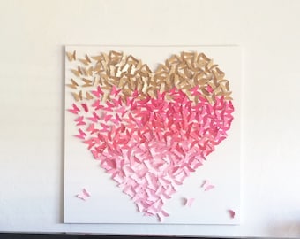 May include: A heart-shaped wall art piece made of paper butterflies in shades of pink and gold. The butterflies are arranged in a gradient pattern, with the darkest pink at the center and the lightest pink at the edges. The butterflies are all facing the same direction, creating a sense of movement.