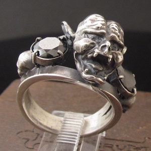 Puede incluir: Un anillo de plata con una piedra preciosa negra y una escultura de plata detallada de una cara grotesca.