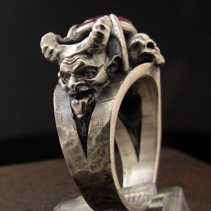 May include: A silver ring featuring an intricate design of a demon's head with horns and a red gemstone. The ring has a textured band.