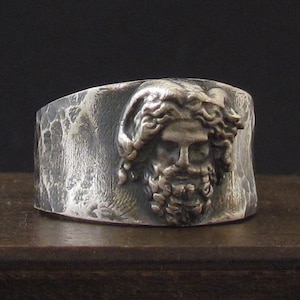 May include: A silver ring with a textured band and a detailed sculpted face of a man with a beard and long hair.