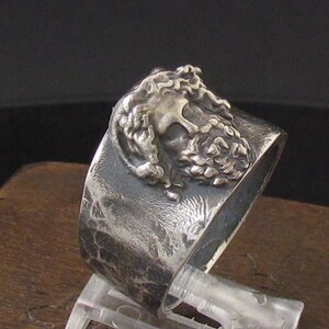 May include: A silver ring with a textured band and a detailed, three-dimensional design of a man's face.