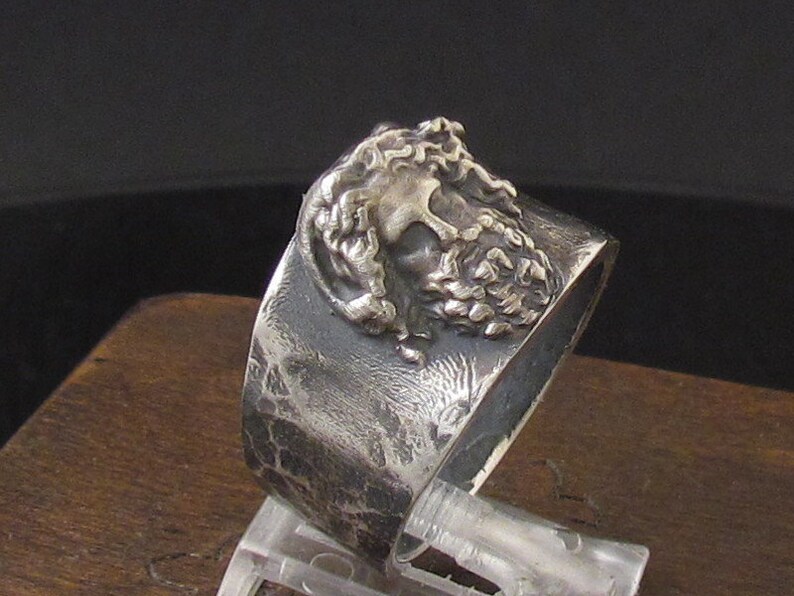 May include: A silver ring with a textured band and a detailed, three-dimensional design of a man's face.