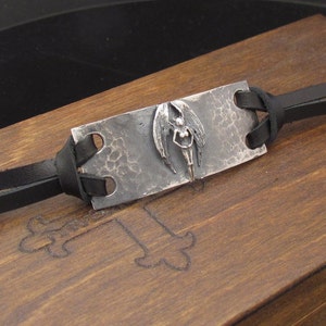 May include: A silver bracelet with a hammered metal plate featuring a detailed angel design. The bracelet is made with black leather straps.