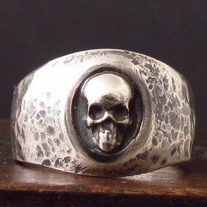 Pu&ograve; includere: Un anello d'argento con una fascia testurizzata e un design a forma di teschio al centro.