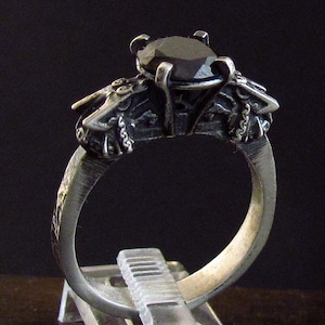 Puede incluir: Un anillo de plata con una gema oscura. El anillo presenta detalles intrincados alrededor del engaste de la gema, con una banda texturizada. La gema oscura está facetada. El anillo se muestra sobre un fondo oscuro.