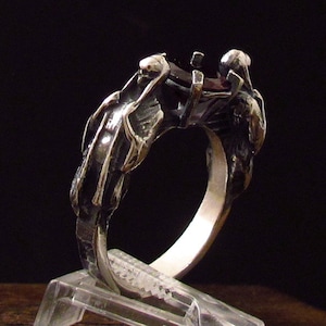 May include: A silver-toned ring with a dark red gemstone setting. The ring features a detailed design of skeletal figures on each side, with intricate carvings and a polished inner band. The ring is displayed on a clear stand against a dark background.