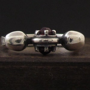 May include: A silver ring with a dark red gemstone set in a geometric design. The ring has two round silver accents on either side of the gemstone.