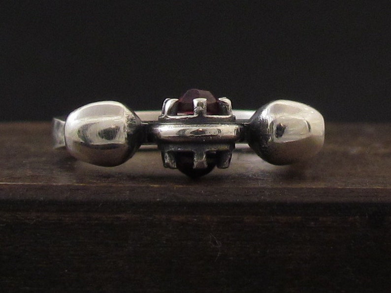 May include: A silver ring with a dark red gemstone set in a geometric design. The ring has two round silver accents on either side of the gemstone.