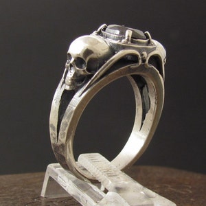 Puede incluir: Un anillo de plata con una piedra preciosa negra y dos detalles de calavera. El anillo está en un expositor transparente.