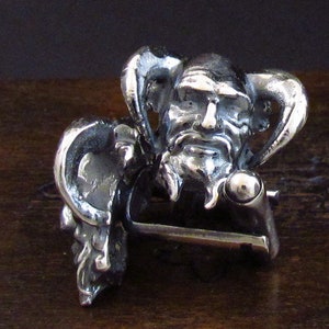 May include: A silver belt buckle with a detailed design of a jester's head with horns and a beard.
