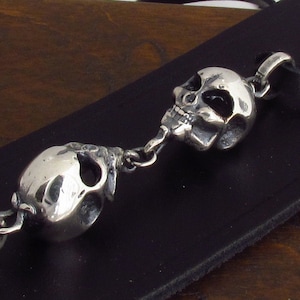 May include: A silver chain with two skull charms. The skulls are detailed and have a realistic look. The chain is attached to a black leather strap.