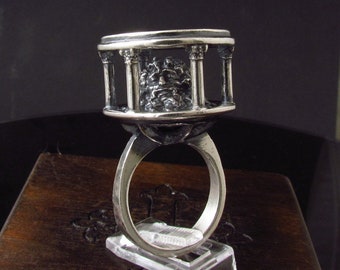 May include: A silver ring with a detailed design of a building with columns and a small figure inside. The ring is on a wooden surface.