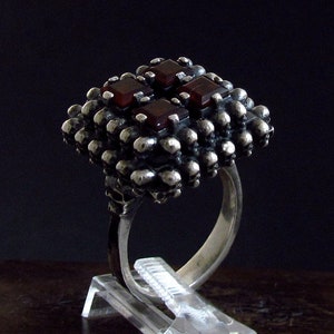 May include: A silver ring with a square-shaped setting featuring multiple red gemstones. The ring is set on a clear display stand.