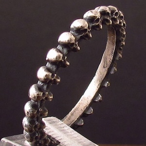 Può includere: Un anello d'argento con un motivo ripetuto di piccoli teschi. I teschi hanno superfici strutturate e sono disposti attorno al gambo dell'anello. L'anello è mostrato su uno sfondo scuro.