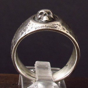 Pu&ograve; includere: Un anello d'argento con un design a forma di teschio sulla parte superiore. L'anello ha una texture martellata.