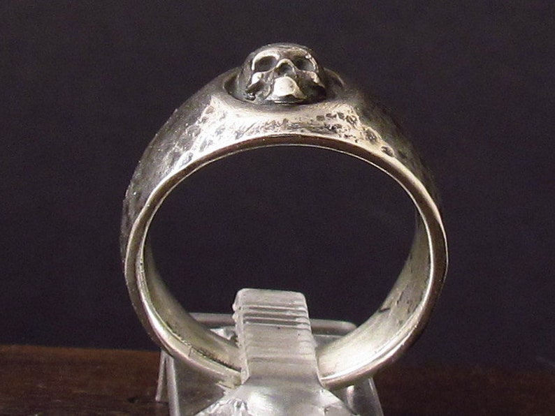 Pu&ograve; includere: Un anello d'argento con un design a forma di teschio sulla parte superiore. L'anello ha una texture martellata.