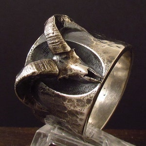 Può includere: Un anello d'argento con una finitura testurizzata e martellata. L'anello presenta un design stilizzato di testa di ariete con corna che si avvolgono attorno alla fascia.