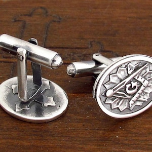 May include: Silver cufflinks with a Masonic symbol design. The symbol features a compass, square, and letter G.