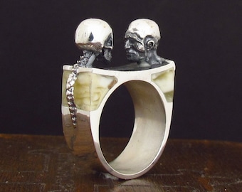 May include: A silver ring with two skulls facing each other. The ring is set with white bone-like material.