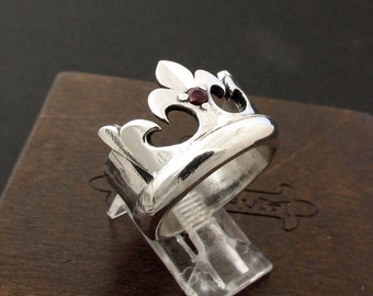 May include: A silver ring with a crown design and a red gemstone in the center.