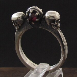 May include: A silver ring with three skull heads and a red gemstone in the center.