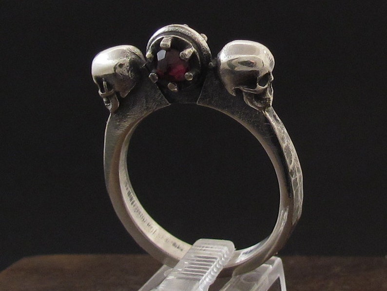 May include: A silver ring with three skull heads and a red gemstone in the center.