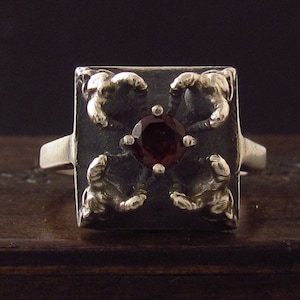 May include: A silver ring with a square face featuring a dark red gemstone surrounded by four stylized silver claws.