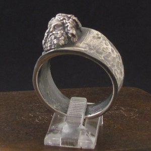 May include: A silver ring with a textured band and a detailed face sculpture on the top.