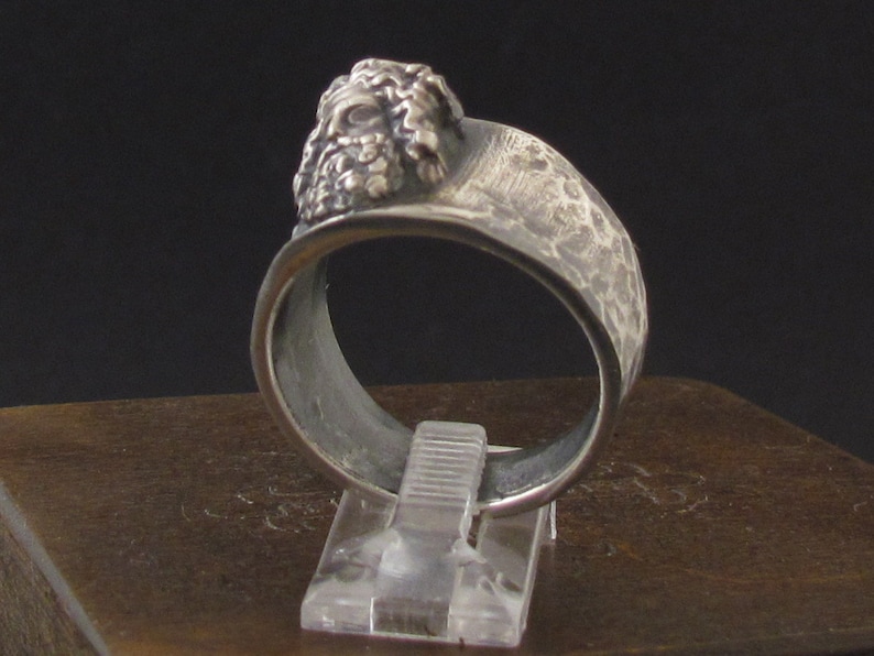 May include: A silver ring with a textured band and a detailed face sculpture on the top.