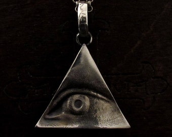 Peut inclure: Un pendentif en argent en forme de triangle avec un œil au centre. L'œil est détaillé avec une pupille et un iris.