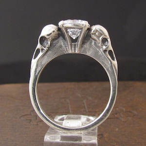 May include: A silver ring with a large, clear gemstone set in a prong setting. The ring is decorated with two skull-shaped accents on either side of the gemstone.