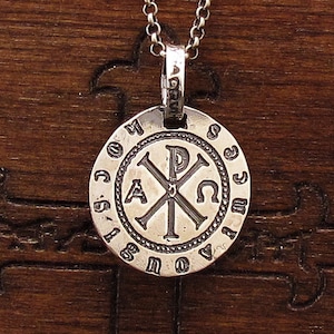 May include: Silver pendant necklace with a round charm featuring a cross with the Greek letters Alpha and Omega, surrounded by a circle of Greek letters.