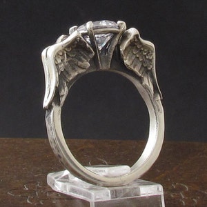 Könnte beinhalten: Ein silberner Ring mit zwei Flügeln auf jeder Seite eines klaren Edelsteins. Die Flügel sind detailliert und haben eine strukturierte Oberfläche.