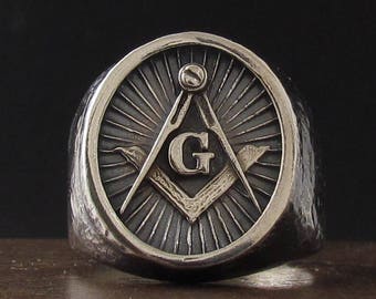 May include: A silver ring with a Masonic symbol, featuring a compass and square with a 'G' in the center, surrounded by a radiating sunburst design.