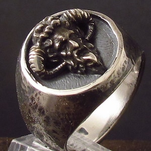 May include: A silver ring with a detailed, textured design of a ram's head with horns. The ring has a hammered finish and a rounded band.