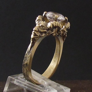 May include: A gold ring with a large clear gemstone set in a detailed, textured design. The ring is made of brass and has a unique, organic shape.