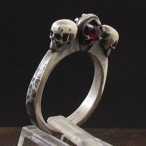 May include: A silver ring with three skull-shaped accents and a red gemstone in the center.