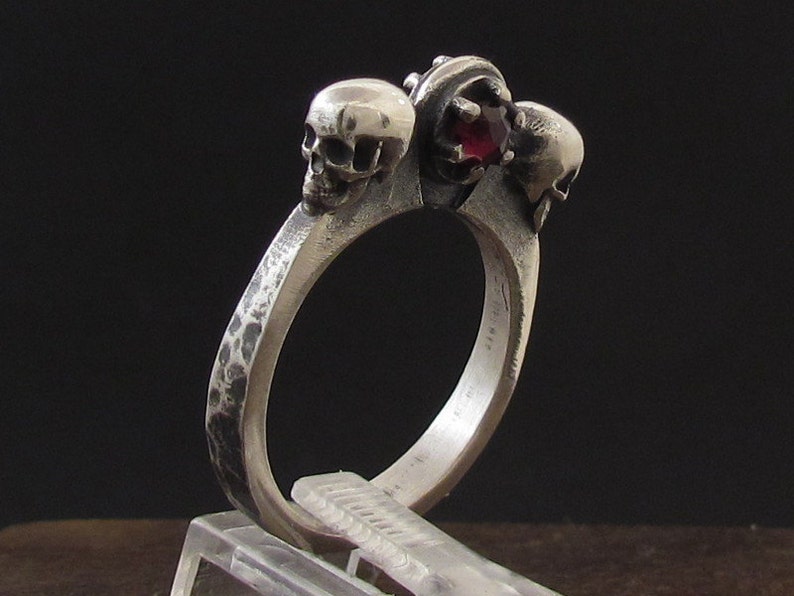 May include: A silver ring with three skull-shaped accents and a red gemstone in the center.