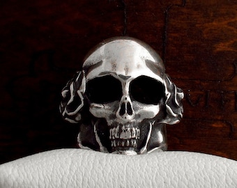 May include: A silver skull ring with detailed floral designs on the sides. The skull has a toothy grin and dark eye sockets.