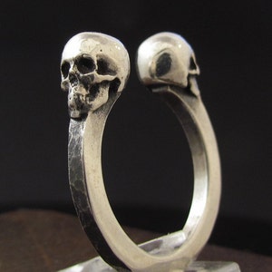 May include: A silver ring with two skull heads facing opposite directions. The ring is made of a thick band of metal with a hammered texture.
