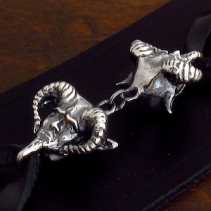 May include: Two silver-toned, detailed, ram skull charms with horns, connected by a silver chain. The charms are on a black leather strap.