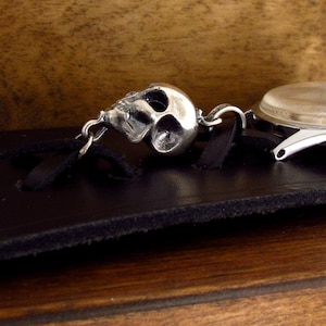 May include: A silver skull charm attached to a watch strap with a black leather band.