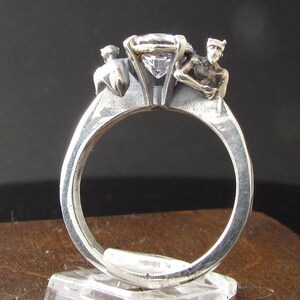May include: A silver ring with a large clear gemstone and two small silver figures on either side of the stone.