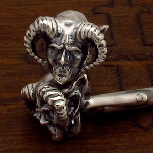 May include: Silver cufflinks featuring a detailed sculpture of a horned demon with a menacing expression.