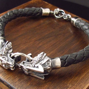 May include: A black leather braided bracelet with silver-toned metal clasps featuring a detailed design. The clasps are shaped like animal heads.