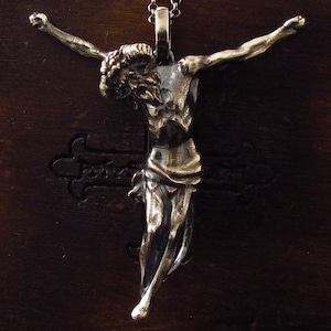 Può includere: Un ciondolo d'argento che raffigura una scultura dettagliata di una persona con le braccia distese. Il ciondolo è appeso a una catena d'argento su uno sfondo di legno marrone scuro.