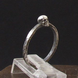 Può includere: Un anello d'argento con un piccolo design a forma di teschio sulla parte superiore. L'anello è realizzato in metallo testurizzato e ha uno stile semplice e minimalista.